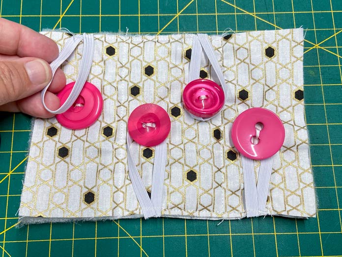 A hand holds a loop of braided elastic sewn to the edge of a quilt sandwich of white, black, and gold fabric on top of a green cutting mat. 3 additional elastic loops and 4 pink buttons have been sewn onto the same quilt square. UNIQUE SEWING Self-Gripping Fastener Strip - 25cm (10″) - White, UNIQUE Webbing 25mm x 10m (1″ x 11yd) - White, ELAN Trench Buckle - 45mm (13⁄4″) - Brown, UNIQUE CREATIV Trim Pack - Black & White, UNIQUE SEWING Invisible Magnetic Snaps - Sew-On - 12mm (1⁄2″) - 2 sets,  SOFTKUT Pinking Shears (Scissors) - 81⁄2″ (21.6cm), COSTUMAKERS General Purpose Closed End Zipper 20cm (8″) - White - 1700, UNIQUE CREATIV Trim Pack - Red & Green, OLFA RTY-2/DX - Deluxe Ergonomic Handle Rotary Cutter 45mm, UNIQUE QUILTING Clever Clips Small - 12 pcs, UNIQUE SEWING Double Adjust Parachute Buckle - Plastic - 25mm (1″) - White, CRAFTING ESSENTIALS Bottle of Buttons - Pink Tones - 75g (2.6oz), UNIQUE Braided Elastic 3mm x 3.7m - White, free tutorial