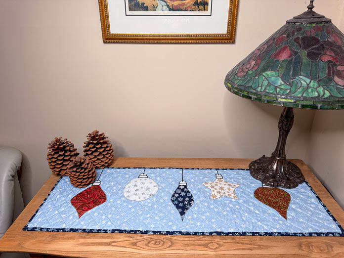 A table runner with 5 Christmas decorations appliqued on a blue snowflake fabric. The table runner is on an antique dresser flanked by three very large pinecones on the left side and a Tyffany-style, stained glass lamp on the right; Gütermann Dekor Metallic Thread 200m – Gold, Gütermann Dekor Metallic Thread 200m - Col. 41, Gütermann Dekor Metallic Thread 200m – Red, Gütermann Dekor Metallic Thread 200m - Christmas Green, Gütermann Dekor Metallic Thread 200m - Col. 1005, Gütermann Denim Thread 100m – Black, UNIQUE Baby Rick Rack 3mm x 3.7m – Scarlet, UNIQUE Medium Rick Rack 6mm x 2.3m - Emerald Green, UNIQUE Jumbo Rick Rack 10mm x 2.3m - Metallic Gold, SCHMETZ #1743 Metallic Needles Carded - 80/12 - 5 count, SCHMETZ #1752 Metallic Needles Carded - 90/14 - 5 count, LDH 8½“ Craft Scissors – Purple, HeatnBond Lite Iron-On Adhesive - 43cm x 30m (17″ x 32 ¾yds) bolt, HeatnBond Stitch n Sew Tear-Away Stabilizer - 50.8cm x 22.8m (20″ x 25yds), oliso M3Pro Project Iron – Coral, OLFA 45mm RTY-2/DX/MAG Ergonomic Rotary Cutter – Magenta, UNIQUE Quilting Wool Pressing Mat - 18″ x 24″ - Grey