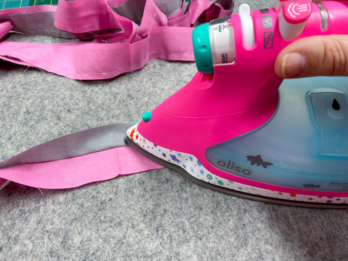 A hand uses a pink iron to press the seam between pink and gray fabric binding strips on a mottled gray wool pressing mat. A pile of pink and gray binding strips can be seen in the background. 