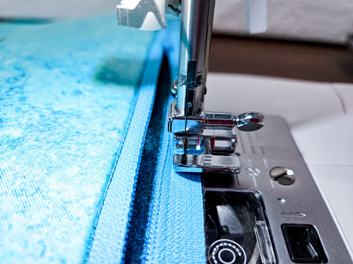 A blue zipper under a metal presser foot on a sewing machine