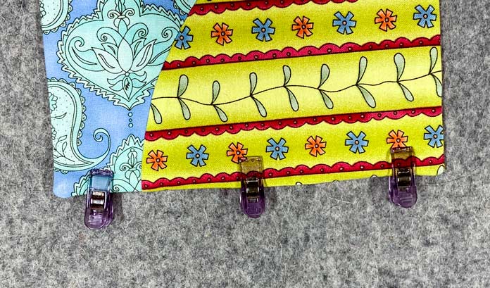 A multicolored quilt block sits on a grey pressing mat. Three purple clips are clipped to the edge.
