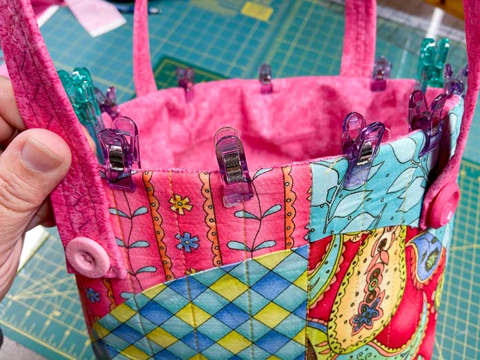 A hand holds one of the handles of a multicolored tote bag. The top edge of the tote bag has small purple clips and large green clips holding the pink lining in place along to folded edge. A green cutting mat can be seen in the background.