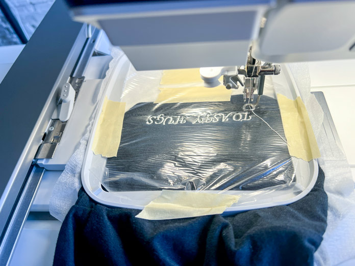 The sleeve is securely framed in a 120x120 hoop, stabilized with white stabilizer on the back, and covered with a layer of clear, water-soluble topping. The sleeve fabric and topping are held firmly in place with masking tape at all four corners of the hoop to ensure no shifting during stitching. The embroidery is in progress 