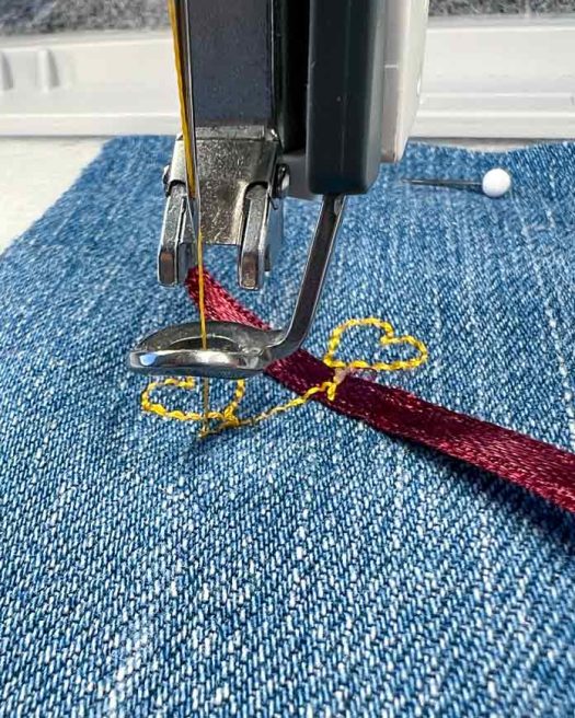 Close-up of the embroidery process using the PFAFF creative icon 2, showing the needle stitching over a satin ribbon placed on denim fabric. The Exclusive Single Ribbon Stitch #8.1.6 is in progress, with the 2A Fancy Stitch Foot guiding the placement. Yellow Mettler 40wt polyester thread is used for the heart motif. The ribbon is aligned with the stitched guide and partially secured under the presser foot as the second heart motif is completed. Background elements include stabilizer beneath the denim and a fabric pin to secure the setup.