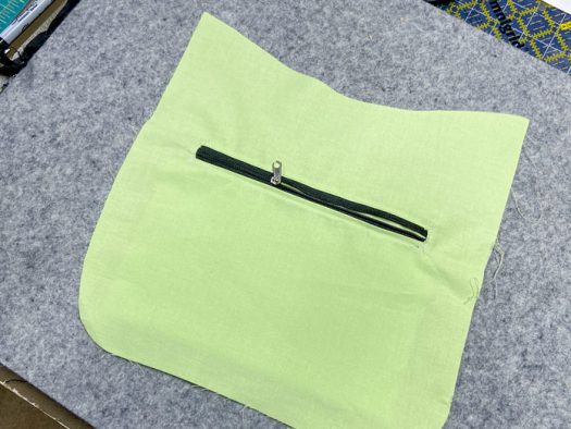 A piece of green lining fabric with an inset black zipper sits atop a grey pressing mat.