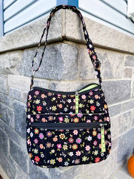 A black tote bag hangs from its strap against the corner of a grey brick building. The purse fabric features tiny flowers throughout and green ribbon zipper pulls.