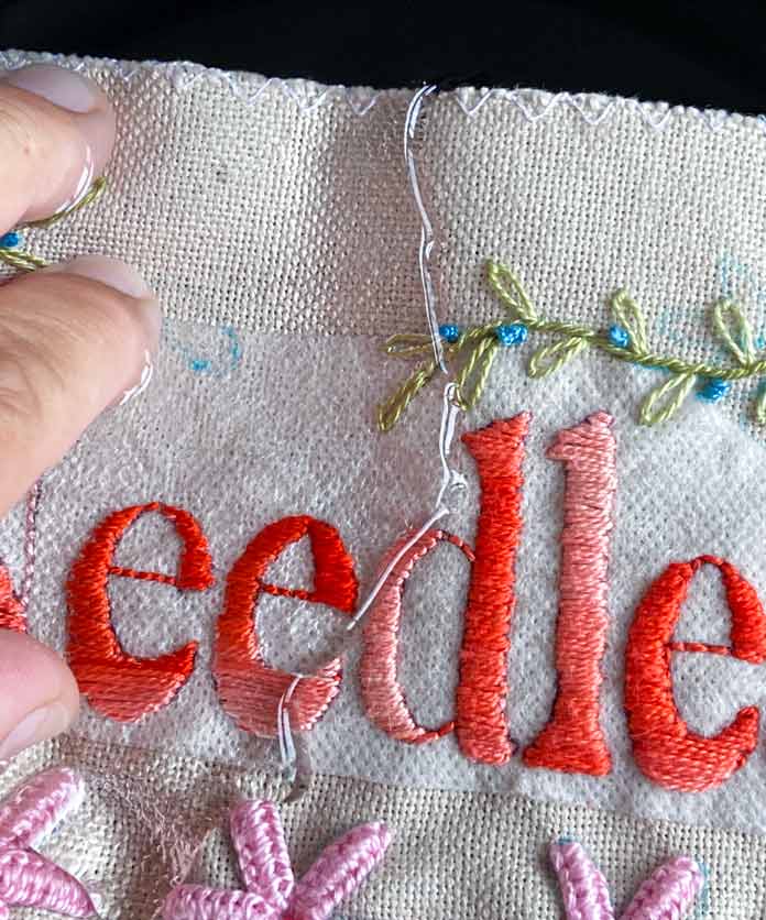 A close-up of a hand holding down a piece of embroidery on beige evenweave fabric soaked in a black bowl of water. The fabric has part of the word ‘Needle’ stitched in a coral-colored thread, parts of 3 pink flowers and a vine of green thread with small blue French knots; MC Charles Craft Monaco Needlework Fabric 28ct 20" x 24" - Tea Dyed, DMC Magic Paper, DMC Embroidery Floss, DMC Cotton Perle Thread, DMC Satin Floss 8 Pack - Radiant Treasures, DMC Light Effects Floss 6 Pack - Tropical Glow, DMC Pearl Cotton Size 3 (14.6m) Thread, DMC Pearl Cotton Size 8 (80m) Thread Balls, UNIQUE Sewing 2-in-1 Dual-Tip Wash-out/Air Erasable Marking Pen