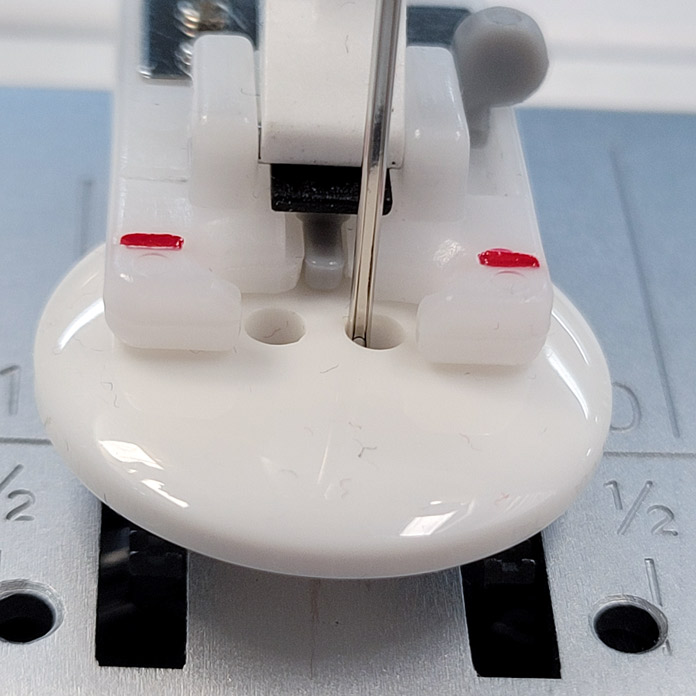 A white button in the slot of a sewing machine presser foot used to sew on buttons; Husqvarna Viking Designer Ruby 90, Button Foot with Placement Tool