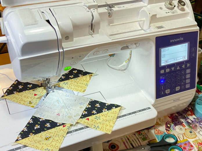 Two pieced black and yellow units for the flying geese block are shown on the bed of the Brother NQ900 sewing machine. Another yellow square is sewn to each of the units.