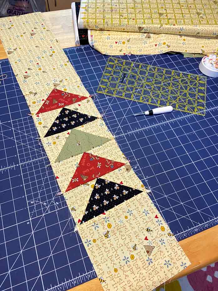 One flying geese border made of yellow border fabric and black, red and green flying geese lays on a blue cutting mat. A seam ripper, rotary cutting ruler, pin cushion and bolt of yellow fabric are also visible; Omnigrid ruler - 6" x 24", OLFA Rotary Cutter, OLFA Double Sided Cutting Mat (navy blue)