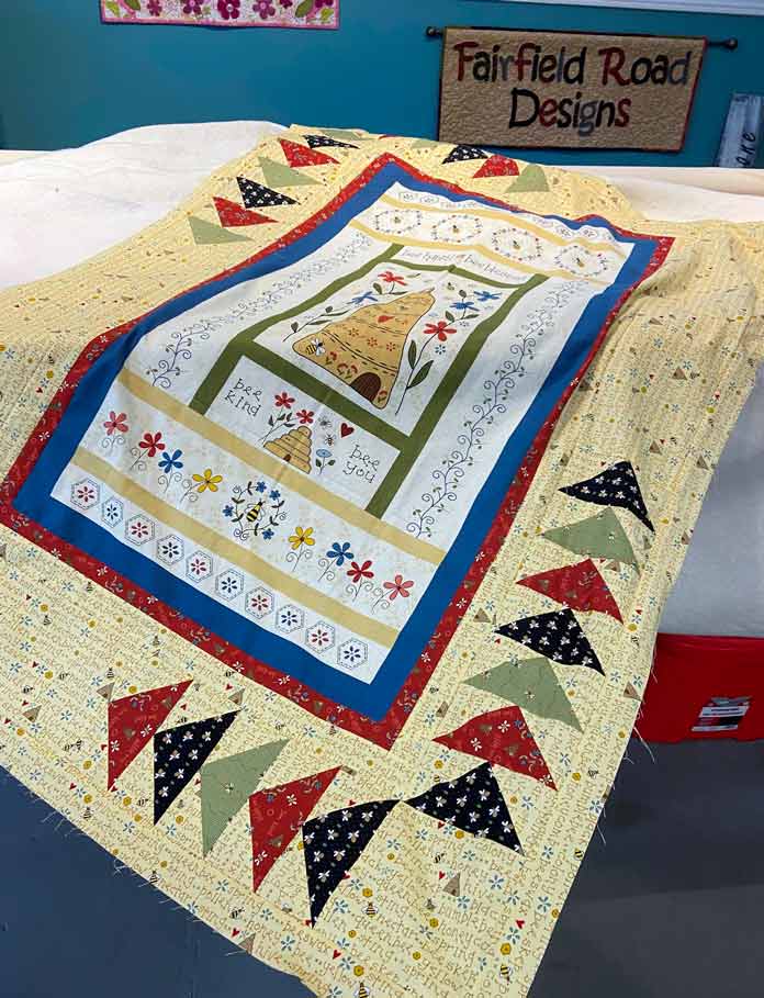 A bee-themed panel with yellow borders and black, green and red flying geese borders lays on the bed of a longarm quilting machine.