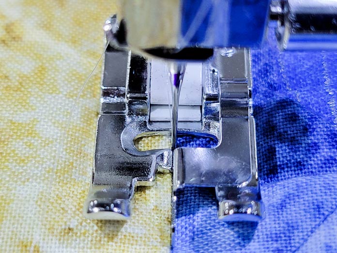 A metal presser foot with a guide in front of beige and blue fabric; Husqvarna Viking Tribute 150C