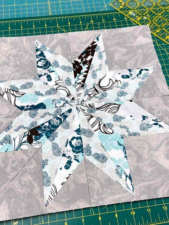 A grey, teal, white and turquoise Star of the East paper-pieced quilt block on a green cutting mat. StitchnSew EZ-Print Quilt Block Sheets, clover roll & Press, Clover Ball Point Awl, Fairfield Crafter’s Choice Pillow Form, Omnigrid 6"x 24" Ruler, OLFA 45mm Deluxe Ergonomic Handle Rotary Cutter, UNIQUE Double Sided cutting Mat