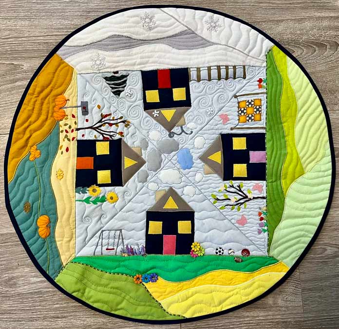 A photo of the completed Circle The Seasons Table Topper with 4 seasons of landscape depicted inside a circle, appliqued, quilted and embellished