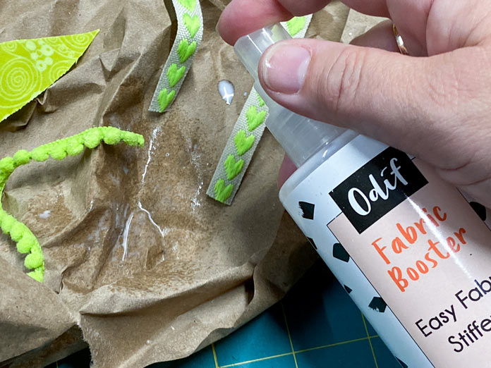 A hand is shown spraying a white liquid onto the end of a green ribbon. Other ribbons and a green fabric leaf can also be seen. A crumpled piece of brown paper is the background; Odif Fabric Booster