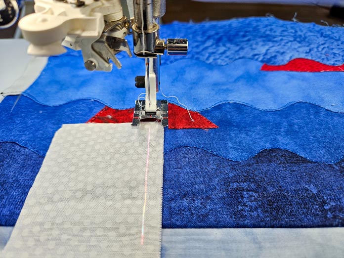 A white fabric printed with small white dots is on a sewing machine with an open-toe sewing machine foot in preparation for stitching to a small quilted scene made from blue and red fabric. A projected light shows a straight line where the machine will stitch. Husqvarna VIKING DESIGNER EPIC 3 sewing and embroidery machine, Husqvarna VIKING 6in/15.2cm Applique Scissor, Husqvarna VIKING Open Toe Presser Foot for IDF System, SULKY Slimline Metallic Dream Assortment