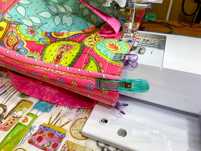 A multicolored tote bag is being sewn by a white sewing machine. There are purple and green plastic clips along the top edge of the bag and a mat with crafting images can be seen under the sewing machine.