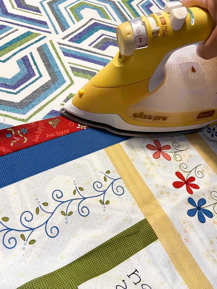 A yellow Oliso Pro iron is shown pressing the seam of a red strip of fabric away from the bee panel on top of a white, teal, blue and gray geometric ironing board. 