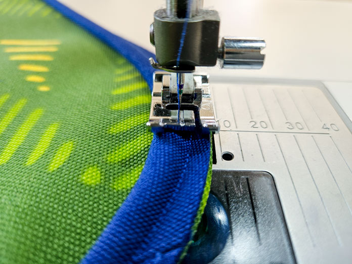 A blue and green fabric under a metal presser foot; Gütermann Nostalgia Box - 50wt Cotton Thread 100m - 48 Shades, Gütermann Nostalgia Box, Flat Felled Foot 9mm, Husqvarna VIKING Opal 690Q, free sewing pattern, outdoor accessories, outdoor cushions, piping; Welt Cord Foot