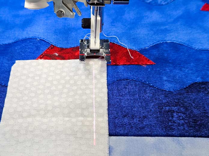 White fabric on top of red and blue fabric under an open-toe sewing machine foot with white thread in a needle in the up position. A red straight line is projected in front of the needle. Husqvarna VIKING DESIGNER EPIC 3 sewing and embroidery machine, Husqvarna VIKING 6in/15.2cm Applique Scissor, Husqvarna VIKING Open Toe Presser Foot for IDF System, SULKY Slimline Metallic Dream Assortment