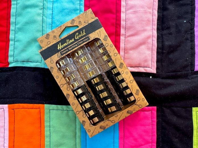 A package of 30 Hemline Gold clips placed on top of a brightly colored strip quilt. Hemline Gold Multi-Use Craft Bag (28.5cm x 36.5cm x 17.8cm), Hemline Gold Sewing Kit, Hemline Gold Quilters 4-in-1 Multi-Mat (30cm x 24cm), Hemline Gold Quilters Clips (Pack of 30), Hemline Gold Retractable Tape Measure - 150cm/60, oliso M3Pro Project Iron – Coral, OLFA 45mm RTY-2/DX/MAG Ergonomic Rotary Cutter – Magenta, OLFA RM-MG - 24″ x 36″ Double Sided Rotary Mat, UNIQUE Sewing Tools Holder, OLFA QR-6x12 - 6″ x 12″ Frosted Acrylic Ruler, OLFA QR-6x24 - 6″ x 24″ Frosted Acrylic Ruler, Fairfield Quilter's 80/20 Quilt Batting - 229 x 274cm (90″ x 108″) 