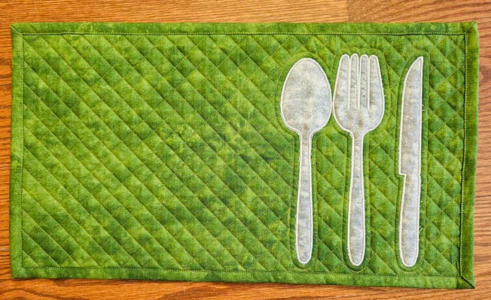 A green placemat with a gray applique spoon, fork, and knife sits on a wooden table. Husqvarna VIKING DESIGNER EPIC 3 sewing and embroidery machine, Husqvarna VIKING 6in/15.2cm Applique Scissor, Husqvarna VIKING Open Toe Presser Foot for IDF System, Husqvarna VIKING Mega Turnable Hoop 450 mm x 450 mm, Husqvarna VIKING Tear-a-Way Extra Wide Stabilizer 24" x 10 Yards, Husqvarna VIKING Cut-a-Way Extra Wide Stabilizer 24” x 10 Yards, SULKY Slimline Metallic Dream Assortment