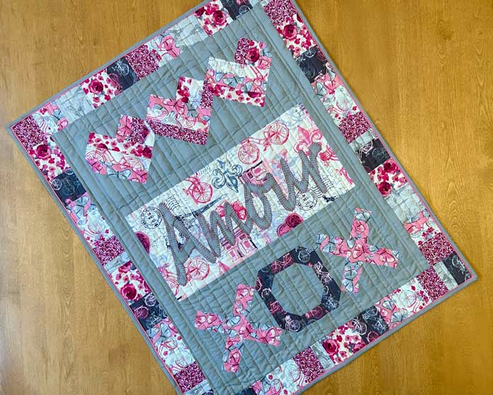 A pink, gray and white quilt with pieced hearts, Amour appliqued on it and the letters XOX along the bottom sits on top of a wooden table.