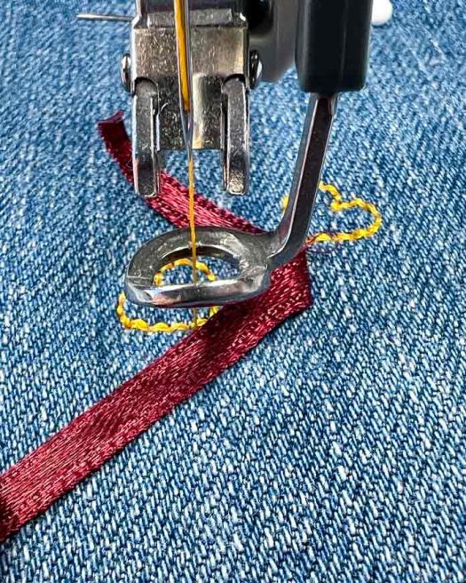 Close-up view of the PFAFF 6D Dynamic Spring Foot stitching over a folded section of 4 mm burgundy satin ribbon on blue denim. The machine is actively completing a heart-shaped motif using a yellow 40wt polyester thread as part of the Single Ribbon Stitch #8.1.6 technique. The ribbon is carefully folded at an angle to align over the previous motif, following the placement guide between stitch repetitions. The fabric is securely hooped, and a white-headed pin is visible in the background for alignment reference.