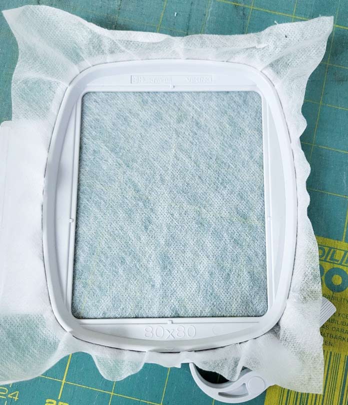 A white non-woven water-soluble stabilizer in an embroidery hoop; Small Square Hoop (80mm x 80mm), Inspira Aqua Magic