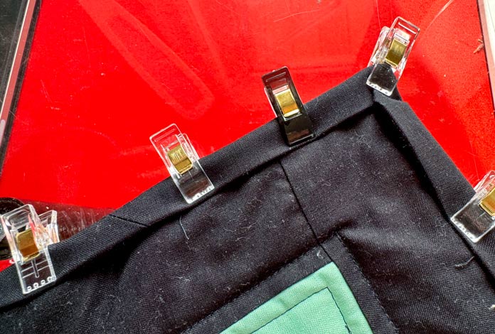 The corner of a black binding is folded to the front corner of a black and green quilt and held in place using Hemline Gold quilt clips with a red sewing table in the background. Hemline Gold Multi-Use Craft Bag (28.5cm x 36.5cm x 17.8cm), Hemline Gold Sewing Kit, Hemline Gold Quilters 4-in-1 Multi-Mat (30cm x 24cm), Hemline Gold Quilters Clips (Pack of 30), Hemline Gold Retractable Tape Measure - 150cm/60, oliso M3Pro Project Iron – Coral, OLFA 45mm RTY-2/DX/MAG Ergonomic Rotary Cutter – Magenta, OLFA RM-MG - 24″ x 36″ Double Sided Rotary Mat, UNIQUE Sewing Tools Holder, OLFA QR-6x12 - 6″ x 12″ Frosted Acrylic Ruler, OLFA QR-6x24 - 6″ x 24″ Frosted Acrylic Ruler, Fairfield Quilter's 80/20 Quilt Batting - 229 x 274cm (90″ x 108″) 