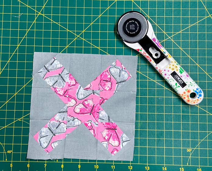 A pink and gray X block and a white rotary cutter with flower decorations on the handle are shown sitting on a green cutting mat.