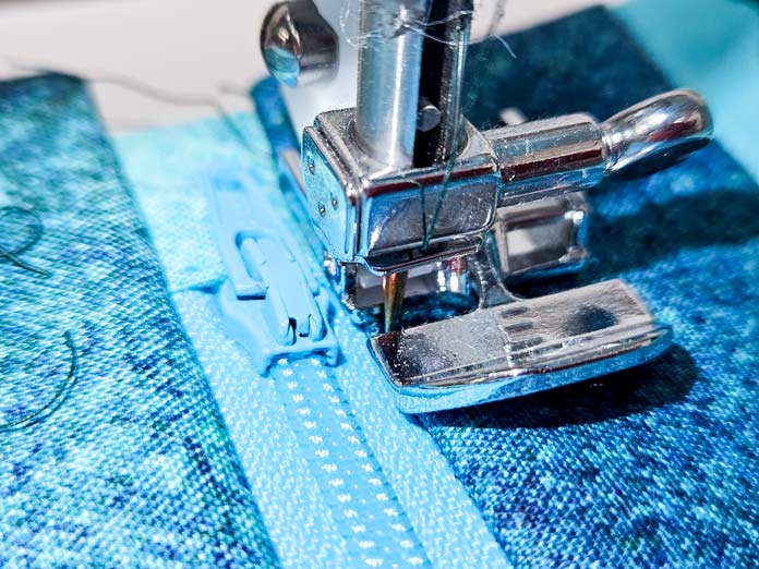A blue zipper with a metal zipper foot on a sewing machine