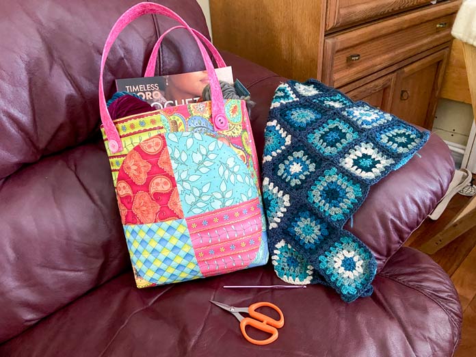 A multicolored tote bag filled with yarn and books sits on a burgundy leather couch with a partially completed blue and white granny square sweater beside it. A crochet hook and pair of orange-handled scissors sit in front of the bag.