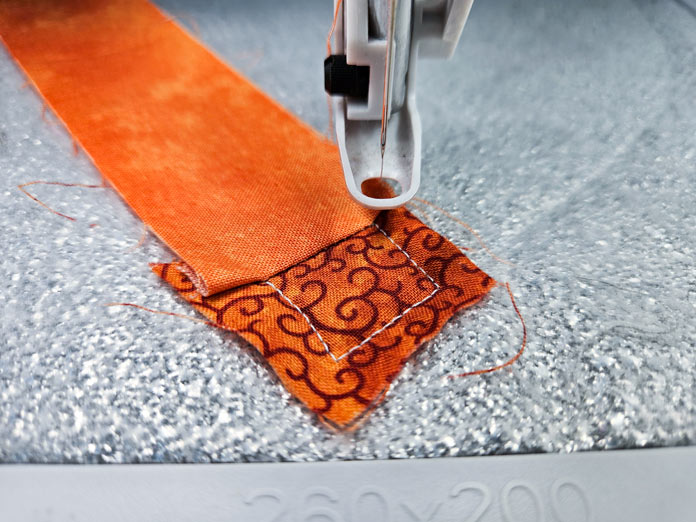 Two pieces of orange fabric on silver fabric and the presser foot of an embroidery machine; Husqvarna VIKING DESIGNER EPIC 3, Husqvarna VIKING Quilters Metal Embroidery Hoop 200mm by 200mm, UNIQUE therm fleece, Whisper Web Mesh Stabilizer