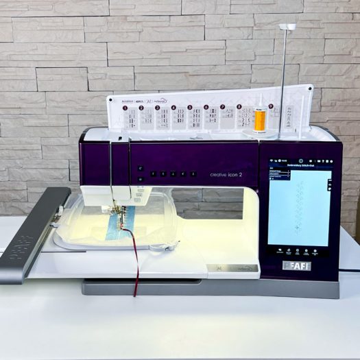 Front view of the PFAFF creative icon 2 set up for embroidery in Stitch-Out mode. The machine is actively stitching a decorative sequence using a folded satin ribbon on a strip of denim fabric, hooped and stabilized in the Elite Hoop 260 x 200 mm. The screen on the right shows the embroidery progress with a design consisting of repeating heart motifs. The 6D Dynamic Spring Foot is engaged, and the selected stitch path is visible in the full-color interface. The upper thread is a 40wt polyester, with the built-in stitch chart and spool holder clearly visible above the machine.