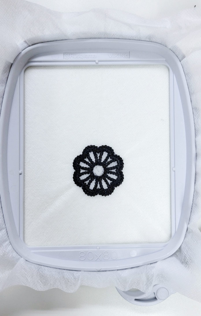 A stand-alone embroidery design in a black thread in a machine embroidery hoop with a water-soluble stabilizer; Small Square Hoop (80mm x 80mm), Inspira Aqua Magic
