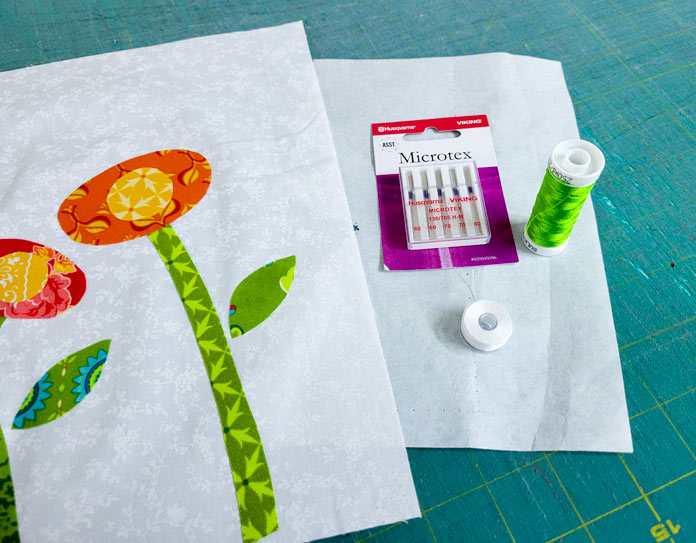 A whimsical flower on a white background, a spool of green thread, a package of needles, and a bobbin of white thread; Inspira Tear-A-Way stabilizer
