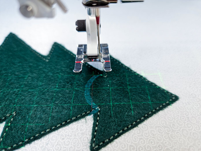 A green Christmas tree on white fabric under an open-toe sewing machine foot with a stitch projection showing where a decorative stitch garland will be stitched on the tree. Husqvarna VIKING DESIGNER EPIC 3 sewing and embroidery machine, Husqvarna VIKING 6in/15.2cm Applique Scissor, Husqvarna VIKING Open Toe Presser Foot for IDF System, SULKY Slimline Metallic Dream Assortment