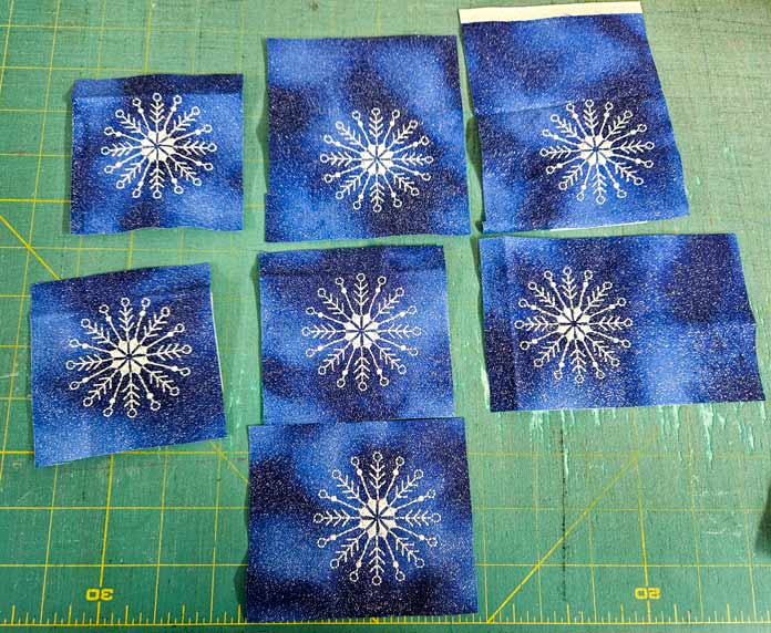 7 white machine embroidery snowflakes on blue fabric sit on top of a green cutting mat; Husqvarna VIKING Designer Quartz 29, mySewnet Machine Embroidery Software, mySewnet Library, Husqvarna VIKING Tear-a-Way Light Stabilizer 15 Inches x 25 Yards