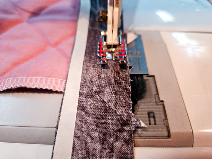 A strip of grey fabric on a pink fabric under a metal presser foot; HeatnBond Lite, quilt binding, fusible web, fabric crafts