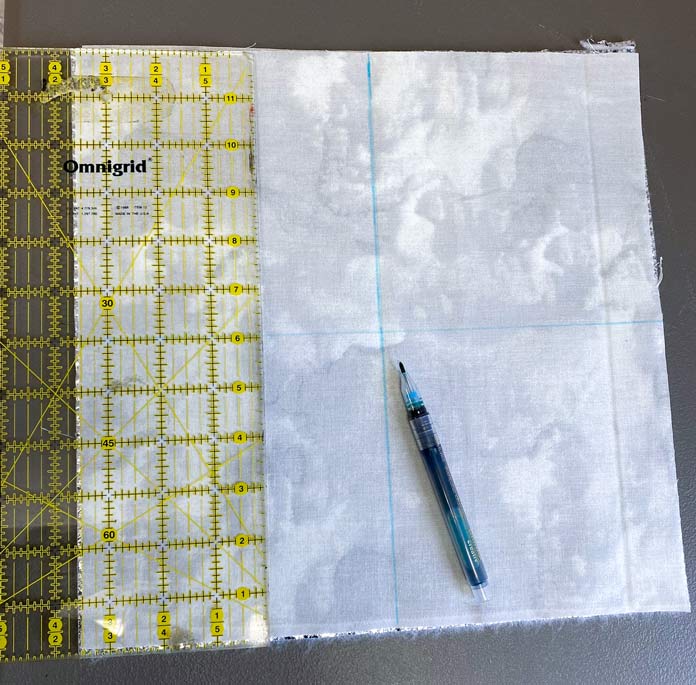 A gray textured piece of fabric with blue lines drawn on it and the pen lying on the fabric next to a quilting ruler; UNIQUE Sewing Wash-out Marker, blue