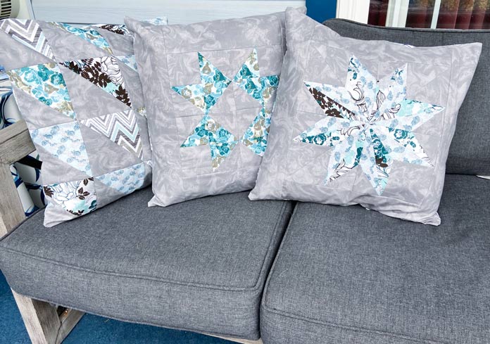 Three grey, white, turquoise and teal cushions sitting on a grey patio couch. Omnigrid Triangle Ruler for Half Square Triangles, Fabric Creations Trendy Neutral Fabric collection, Fabric Creations Fabric Palette collection, Clover Hot Hemmer, UNIQUE quilting Clever Clips, UNIQUE Clear Grip, Oliso Pro TG1600 Smart Iron, Fairfield Crafter’s Choice Pillow Forms, StitchnSew EZ-Print Quilt Block Sheets