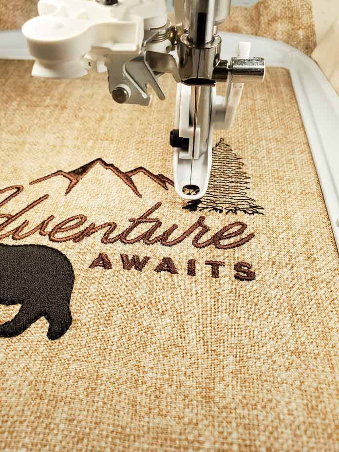 The back of a black bear, brown lettering, and the start of a black tree in a machine embroidery hoop; Husqvarna Viking Designer Ruby 90