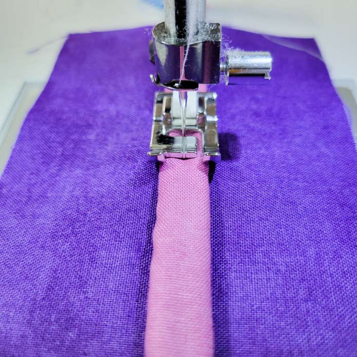 A tube of pink fabric on a purple fabric with a metal presser foot on the sewing machine; Husqvarna Viking Opal 670, Husqvarna Viking Double Welt Cord Foot