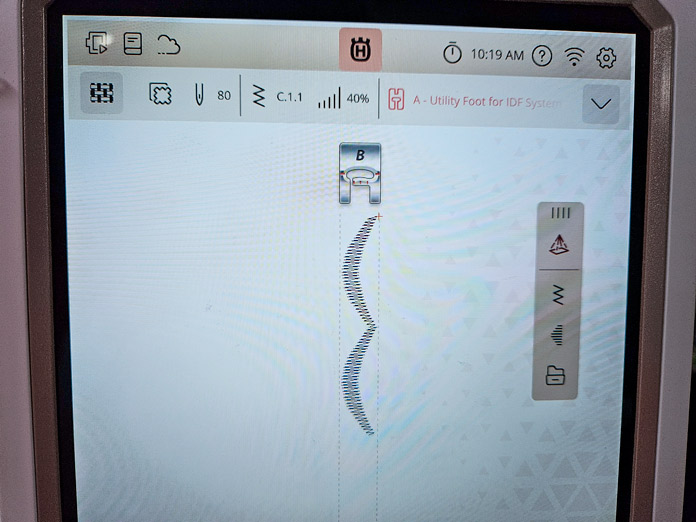 A screen on a computerized sewing machine shows two repeats of a scalloped satin stitch. Husqvarna VIKING DESIGNER EPIC 3 sewing and embroidery machine, Husqvarna VIKING 6in/15.2cm Applique Scissor, Husqvarna VIKING Open Toe Presser Foot for IDF System, SULKY Slimline Metallic Dream Assortment
