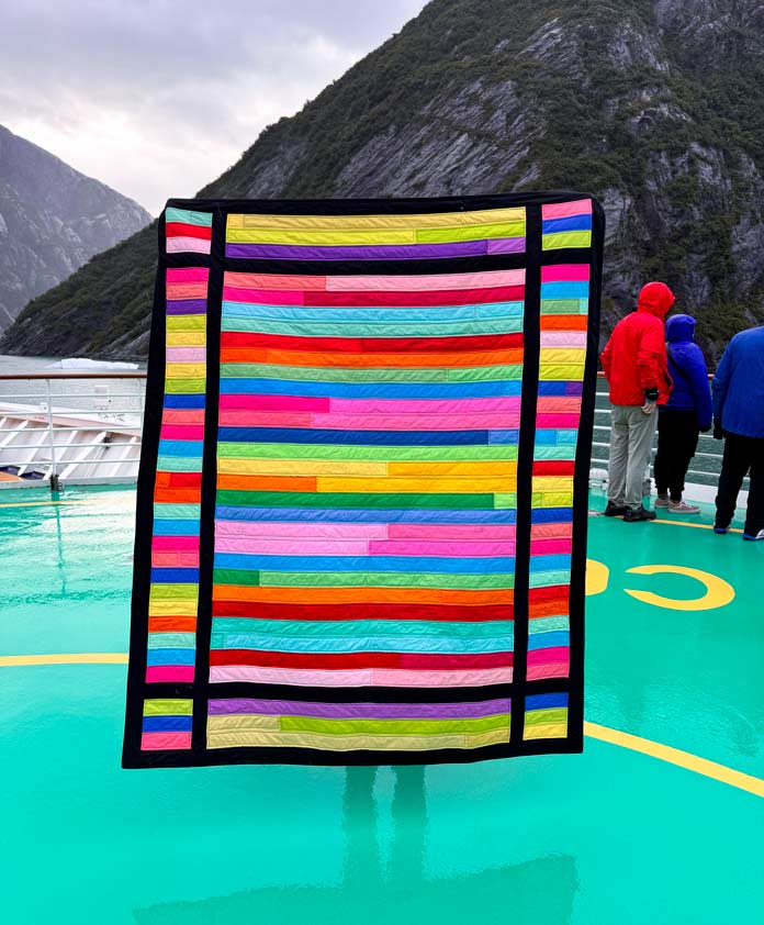 A completed brightly colored strip quilt with the Alaska coast as the background. Hemline Gold Multi-Use Craft Bag (28.5cm x 36.5cm x 17.8cm), Hemline Gold Sewing Kit, Hemline Gold Quilters 4-in-1 Multi-Mat (30cm x 24cm), Hemline Gold Quilters Clips (Pack of 30), Hemline Gold Retractable Tape Measure - 150cm/60, oliso M3Pro Project Iron – Coral, OLFA 45mm RTY-2/DX/MAG Ergonomic Rotary Cutter – Magenta, OLFA RM-MG - 24″ x 36″ Double Sided Rotary Mat, UNIQUE Sewing Tools Holder, OLFA QR-6x12 - 6″ x 12″ Frosted Acrylic Ruler, OLFA QR-6x24 - 6″ x 24″ Frosted Acrylic Ruler, Fairfield Quilter's 80/20 Quilt Batting - 229 x 274cm (90″ x 108″) 