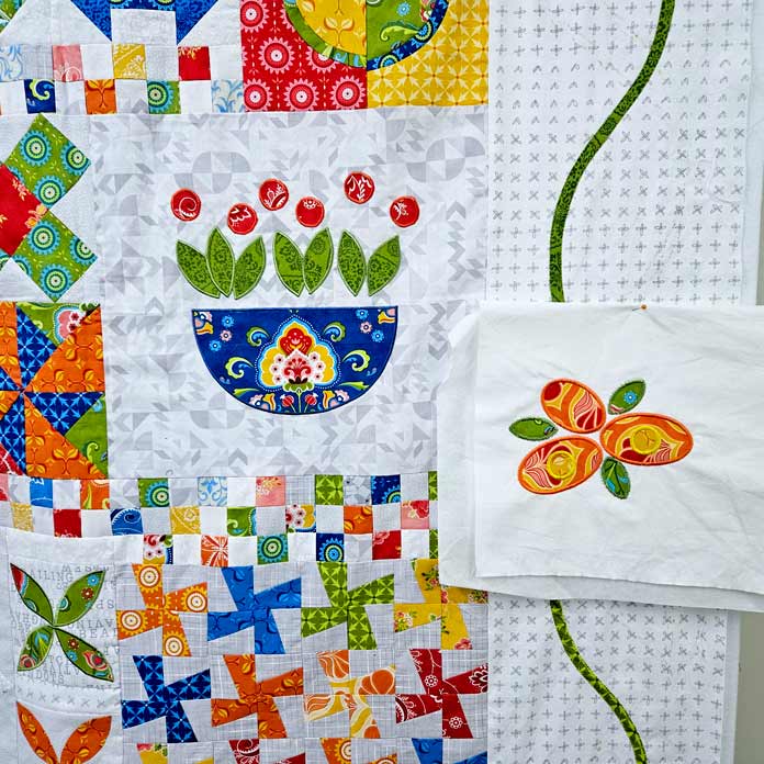 An orange and green floral motif stitched on white fabric next to a colorful quilt