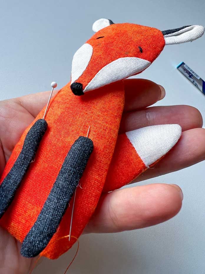 Hand holding a small fabric fox with the head, legs, and tail pinned in place. PFAFF creative icon 2, PFAFF creative icon 2 sewing and embroidery machine, PFAFF Universal Needles Size 80/12, PFAFF Universal Needles Size 90/14, PFAFF Quilting Needles Size 90/14, PFAFF ¼” Quilting Foot for IDT System, Mettler 40wt polyester thread, oliso iron