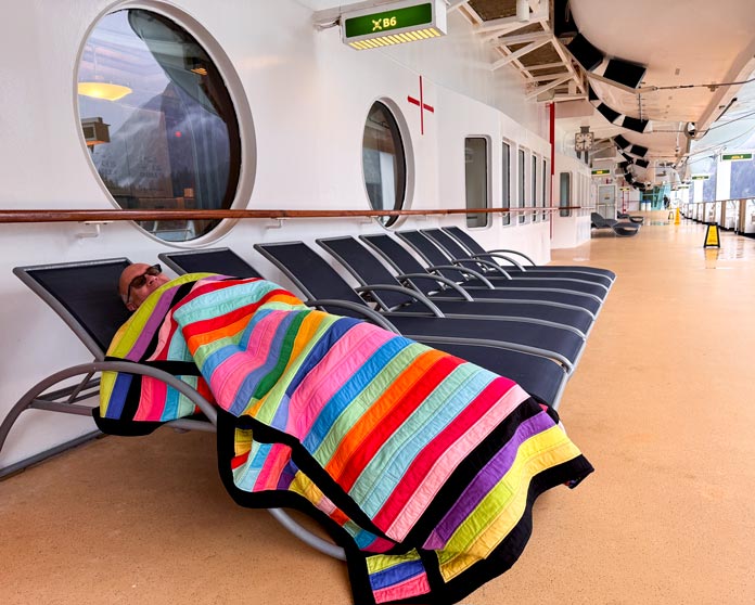 A man lying down covered with a colourful quilt on a lawn chair on a cruise ship in Alaska. Hemline Gold Multi-Use Craft Bag (28.5cm x 36.5cm x 17.8cm), Hemline Gold Sewing Kit, Hemline Gold Quilters 4-in-1 Multi-Mat (30cm x 24cm), Hemline Gold Quilters Clips (Pack of 30), Hemline Gold Retractable Tape Measure - 150cm/60, oliso M3Pro Project Iron – Coral, OLFA 45mm RTY-2/DX/MAG Ergonomic Rotary Cutter – Magenta, OLFA RM-MG - 24″ x 36″ Double Sided Rotary Mat, UNIQUE Sewing Tools Holder, OLFA QR-6x12 - 6″ x 12″ Frosted Acrylic Ruler, OLFA QR-6x24 - 6″ x 24″ Frosted Acrylic Ruler, Fairfield Quilter's 80/20 Quilt Batting - 229 x 274cm (90″ x 108″) 