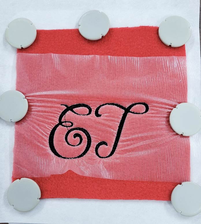 The letters E and T stitched in black thread on red fleece in a metal embroidery hoop with a layer of Inspira Water Works Soluble Film on top; Small Metal Hoop (100m x 100m), Inspira Magnets for Metal Hoop